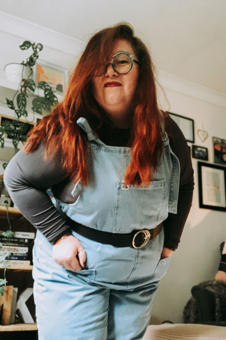 Image of Melanie, she is standing wearing denim dungarees and leaning towards the camera smiling